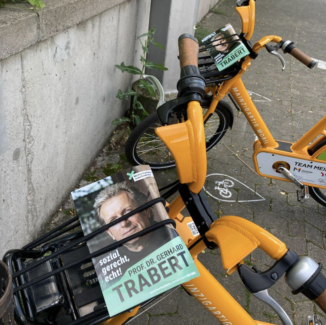 Team Trabert: Zwei gelb-orange Leihfahrräder vor einer Mauer mit Trabert-Flyern auf dem Gepäckständer.